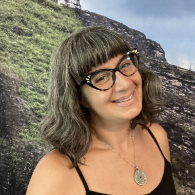 A woman wearing glasses and a flower of life pendant, smiling in front of a grassy knoll