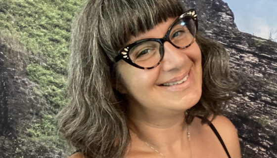 A woman wearing glasses and a flower of life pendant, smiling in front of a grassy knoll