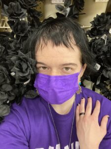 Image description: Selfie of Lucas Scheelk, a white Jew, wearing a purple long-sleeve shirt with a matching purple mask, posing for the camera against a display of fake black roses.