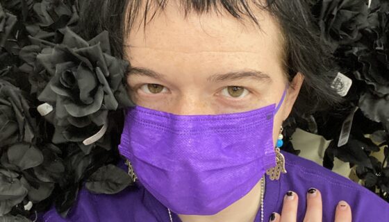 Image description: Selfie of Lucas Scheelk, a white Jew, wearing a purple long-sleeve shirt with a matching purple mask, posing for the camera against a display of fake black roses.